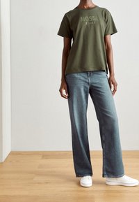 Personne portant un sweat-shirt MOSH vert olive, un jean bleu ample et des baskets blanches, sur un sol en bois avec un fond de mur clair.