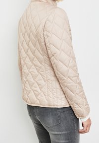 Lys beige vatteret jakke med lynlås, der har et diamantmønster og en høj krave, båret over grå tætsiddende jeans.