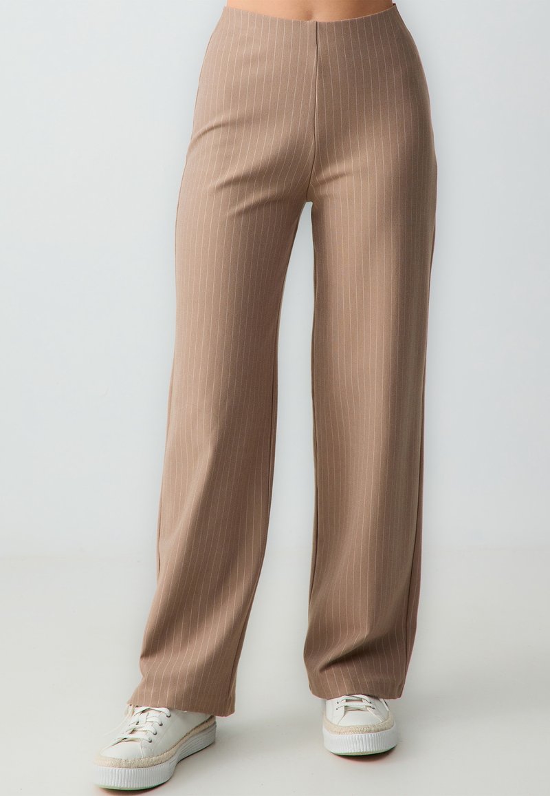 Personne portant un pantalon large à fines rayures beige et des baskets blanches, debout devant un fond uni clair.