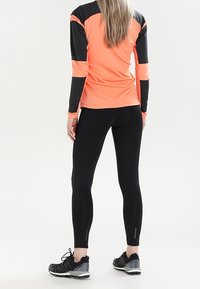 Completo sportivo composto da una maglietta a maniche lunghe arancione brillante con dettagli neri, abbinata a leggings neri e scarpe da corsa grigie.