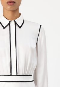 Blouse blanche avec des accents noirs, présentant un devant à boutons, un col classique et une taille froncée. Fabriquée en tissu transparent.