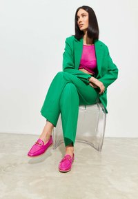 Groen op maat gemaakt pak, roze cropped top en felroze loafers met gouden hardware details. Heldere stoel en neutrale textuurvloer.