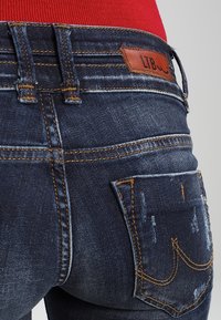 Jeans de mezclilla oscura descoloridos con costuras naranjas, cinco bolsillos y etiqueta de la marca en la cintura trasera. Detalles desgastados en el bolsillo trasero.