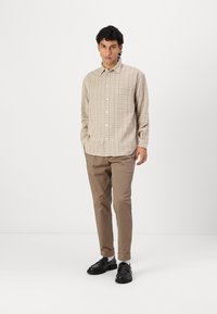 Camisa a cuadros en marrón claro con cierre de botones en la parte delantera y un bolsillo en el pecho, combinada con chinos beige y zapatos negros.