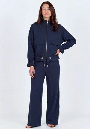 Kvinde iført navy lynlåstrøje med store forlommer og matchende vidde bukser med hvide sidelister, stående mod en ensfarvet baggrund.