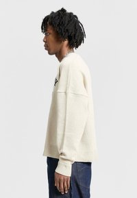 Cream-colored sweater with a textured knit, oversized fit, ribbed cuffs, and hem. Features black text on the front. Worn with dark blue pants.