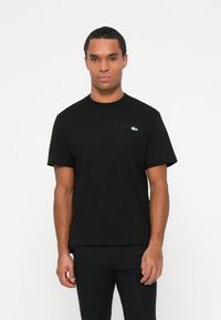 T-shirt en coton noir avec un col rond, des manches courtes et un logo brodé vert discret sur la poitrine gauche. Texture lisse.