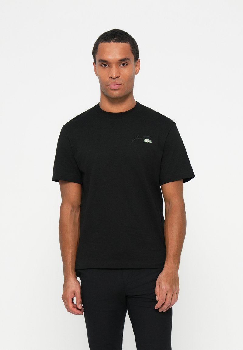 T-shirt en coton noir avec un col rond, des manches courtes et un logo brodé vert discret sur la poitrine gauche. Texture lisse.