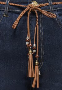 Ceinture en cuir marron tressée nouée en un nœud avec des glands perlés sur un jean en denim bleu foncé, maintenue par un bouton.