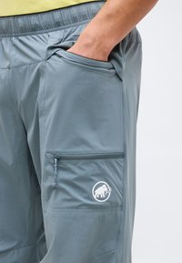 Χέρι στην τσέπη μπλε-γκρι παντελονιού με ελαστική μέση και πλευρική τσέπη με φερμουάρ που φέρει λευκό λογότυπο μαμούθ.