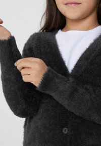 Tmavě šedý huňatý kardigan s výstřihem do V, zapínáním na knoflíky a přiléhavými rukávy. Textura je měkká a načechraná, což zvýrazňuje vizuální teplo.