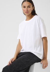 T-shirt en coton blanc avec une coupe décontractée, manches courtes et un logo en forme de cœur discret sur la poitrine, associé à un pantalon texturé sombre.