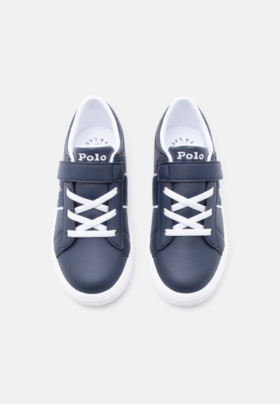 Paire de baskets bleu marine avec semelles blanches, lacets blancs croisés et une bride Velcro, portant la marque "Polo" sur la languette.