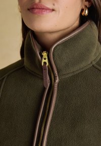 Woman wearing an olive green fleece jacket with brown leather trim and gold zipper, partially zipped to the collar.