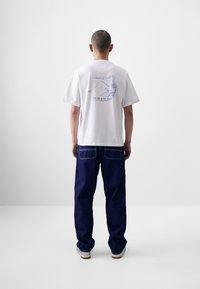 T-shirt branca de algodão com gráfico azul nas costas, combinada com jeans de corte largo azul escuro e sapatos brancos com sola castanho claro.