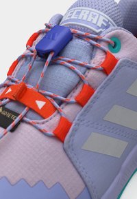 Κοντινό πλάνο μωβ και πορτοκαλί κορδονιών αθλητικού παπουτσιού με μπλε κούμπωμα, που αναδεικνύει την ετικέτα Gore-Tex και το υφασμάτινο πλεκτό ύφασμα στο επάνω μέρος του παπουτσιού.