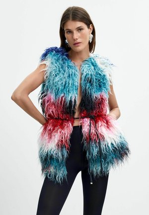 Donna che indossa un gilet soffice multicolore con sfumature di blu, rosso, bianco e nero, abbinato a pantaloni neri, posa con le mani sui fianchi.