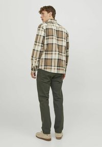 Camisa de franela a cuadros en colores beige, marrón y crema, combinada con pantalones verdes oscuros. La camisa cuenta con un cuello clásico y puños abotonados.