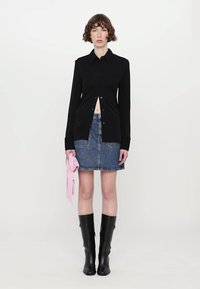 Svart langermet skjorte med knapper og flagrende design, kombinert med et blått denimskjørt. Modellen holder et rosa tilbehør; har på seg svarte støvler til knærne.