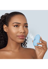Dispositif facial bleu portable, de forme arrondie, avec un bouton unique et un jet de vapeur, tenu par une personne aux cheveux bouclés et à la peau lisse.