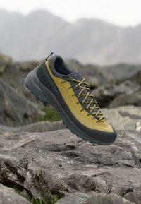 Zapato de senderismo amarillo y gris con cordones verdes flotando sobre un terreno rocoso con un fondo montañoso.