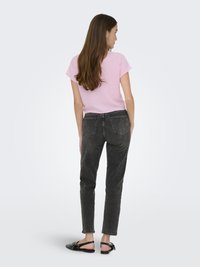 Haut rose à manches courtes, jeans gris taille haute et sandales noires à clous. Cheveux longs, vus de dos sur fond blanc.