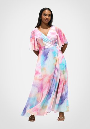 Vrouw draagt een vloeiende, pastelkleurige wikkeljurk met wijde korte mouwen en een split, gecombineerd met sandalen met bandjes op een witte achtergrond.