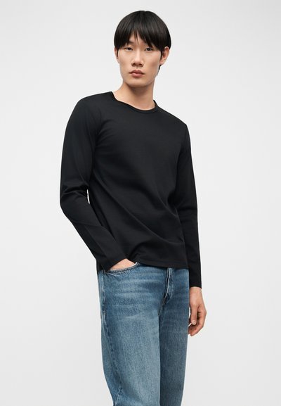 Άνδρας που φοράει μαύρο μακρυμάνικο πουκάμισο και μπλε τζιν, στέκεται με το ένα χέρι στην τσέπη μπροστά σε μια απλή λευκή επιφάνεια.