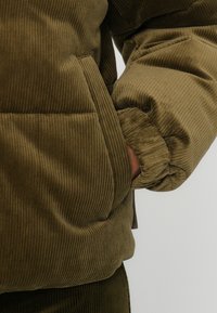 Olivovo zelená zamatová puffer bunda s ryhovanou textúrou, stiahnutými manžetami a bočnými vreckami. Mäkká tkanina s objemným siluetou.