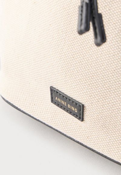 Close-up van een beige tas van gestructureerde stof met zwarte ritstrekker en een zwart label met de tekst "ANINE BING" in gouden letters.