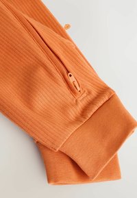 Manche de vêtement orange côtelée avec une poche zippée et un poignet lisse. Le tissu a une texture douce et un motif uniforme.