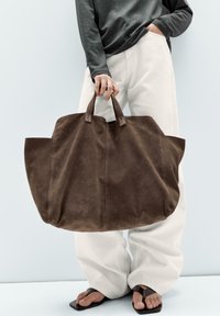 Brun mockaläder toteväska med två robusta handtag, bred öppen design och mjuk textur. Matchad med ljusa, löst sittande byxor.