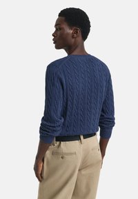 Marineblauwe kabeltrui met een ronde hals, voorzien van geribde mouwen en taille. Gecombineerd met khaki broeken. Geen zichtbare patronen of logo's.