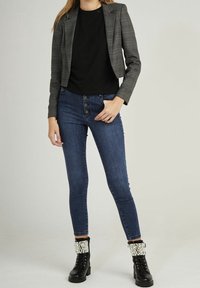 Blazer grigio a quadri, t-shirt nera, jeans blu a vita alta con dettagli dei bottoni, e stivali da combattimento neri con risvolti decorati con perle.
