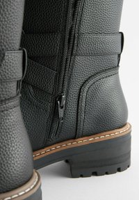Bottine en cuir noir, finition texturée, fermeture éclair sur le côté et couture contrastante marron sur la semelle. Dotée d'un talon carré pour plus de stabilité.