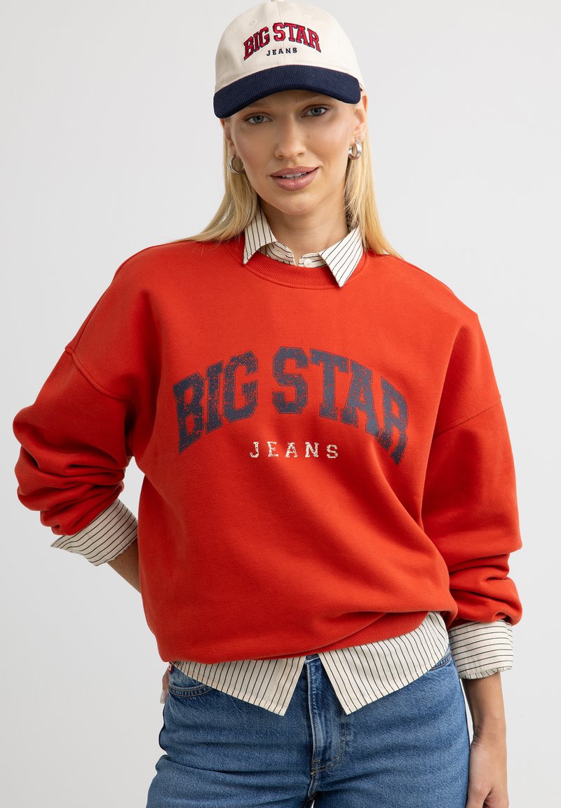 Punane pusakas, millel on tumesinise printimisega "BIG STAR JEANS", koos valge ja tumesinise triibulise kraega särgiga, seljas sinised denimpüksid.