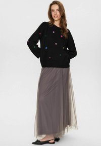 Pull noir avec des pois colorés, coupe ample ; jupe maxi grise transparente, tissu fluide ; chaussures plates noires avec un détail en nœud.