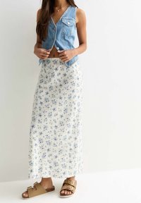 Denim mouwloze vest met knopen aan de voorkant over een witte maxi-jurk met bloemmotief. Bruine sandalen met dubbele kruislings banden.
