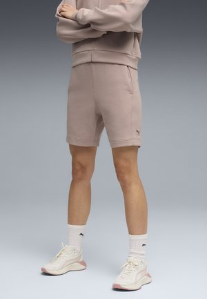 Osoba ubrana w beżowe spodenki Puma i dopasowaną bluzę, białe skarpetki Puma oraz kremowo-różowe sneakersy, stoi ze skrzyżowanymi ramionami.