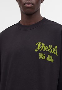 Jeune personne portant un sweat-shirt noir avec le logo Diesel jaune-vert et un texte sur la poitrine.