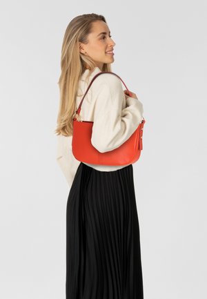 Rode leren handtas met een gestructureerde afwerking, gebogen vorm en een korte schouderband. Draagt bij een beige trui en een zwarte geplooide rok.