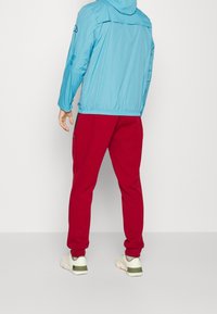 Světle modrá bunda s kapucí a elastickým lemem, červené tepláky s zúženými manžetami a neutrální tenisky se zelenými akcenty.