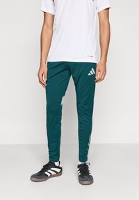 Mėlynos žalsvos sportinės kelnės su trimis baltomis juostelėmis šonuose, pagamintos iš lengvos medžiagos, derinamos su baltu trumpomis rankovėmis marškinėliu ir juodais batais.