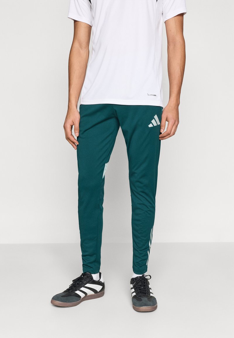 Mėlynos žalsvos sportinės kelnės su trimis baltomis juostelėmis šonuose, pagamintos iš lengvos medžiagos, derinamos su baltu trumpomis rankovėmis marškinėliu ir juodais batais.