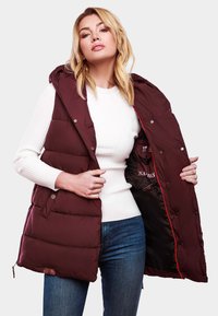 Bordo puffer prsluk s kapuljačom, dugmićima i crvenom unutrašnjošću. Ima kviltani dizajn i logo unutra. Nosio se preko bijele rebraste majice.