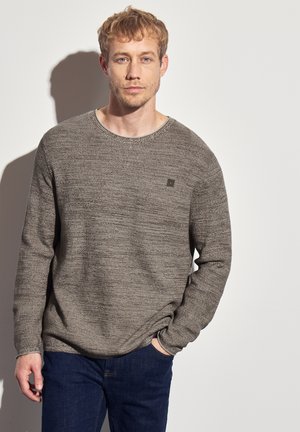 Hellebrauner, strukturierter Strickpullover mit Rundhalsausschnitt und langen Ärmeln, versehen mit einem kleinen Logo auf der Brust. Kombiniert mit dunkelblauen Jeans.