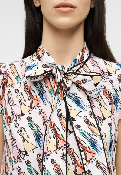 Kvinde iført en bluse med en stor sløjfe ved halsen, med et farverigt print af forskellige modetegninger på hvidt stof.