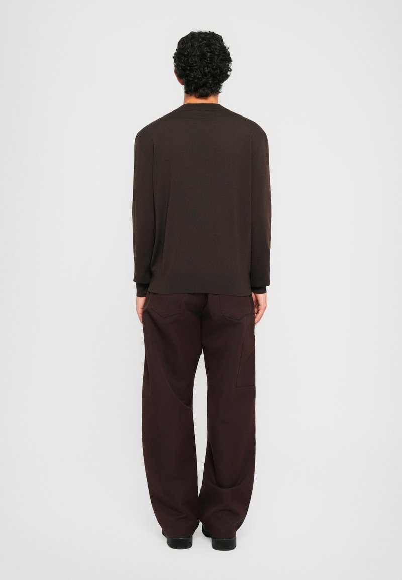 Un homme aux cheveux bouclés se tenant de dos, portant un pull marron foncé et un pantalon marron foncé ample, sur un fond clair et uni.