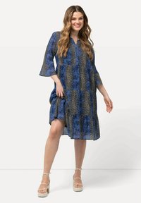 Sininen printtipuku, jossa on kellohihat, kerroksellinen muotoilu ja sekoitus abstrakteja kuvioita. Varustettu kauluksen kanssa ja virtaavasta kankaasta.