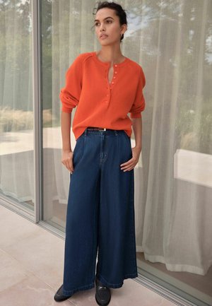 Oranger gestrickter Pullover mit Rundhalsausschnitt und Knopfverschluss, kombiniert mit weit geschnittenen dunklen Denim-Jeans und schwarzen Slippers.